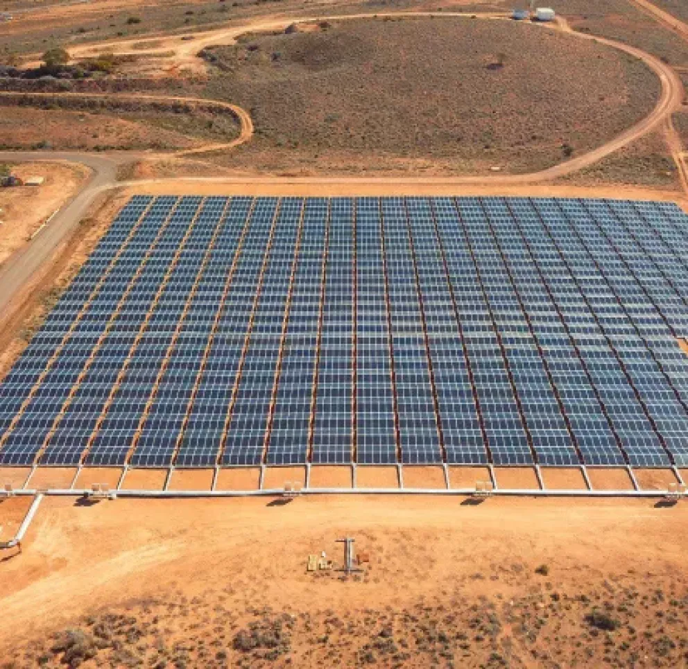 Aerial view of a large solar farm with numerous solar panels arranged in rows on a dry, barren landscape. Dirt roads surround the solar farm, and a small building is visible in the foreground.