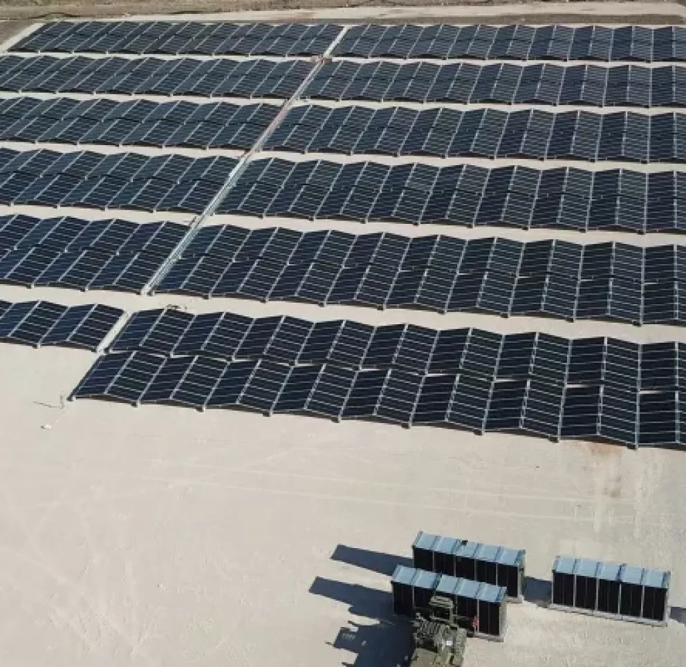 Aerial view of a large solar farm with rows of solar panels on a flat, sandy surface. The panels are arranged in a grid pattern, capturing sunlight for energy production. The surrounding area includes some construction equipment and barren land.