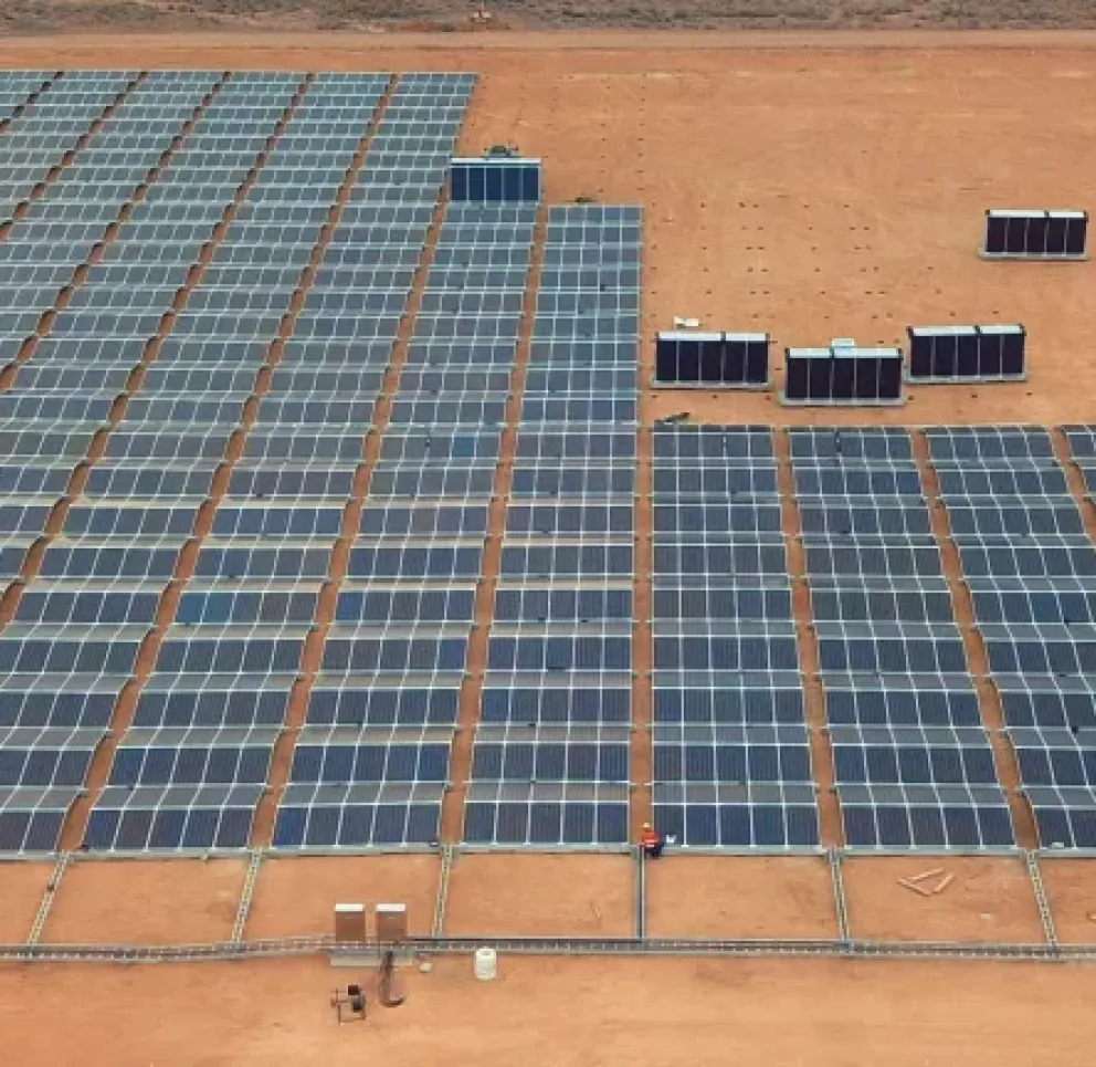 Aerial view of a large solar panel farm on a barren landscape. Rows of solar panels are aligned on reddish-brown soil, with several rectangular structures nearby.