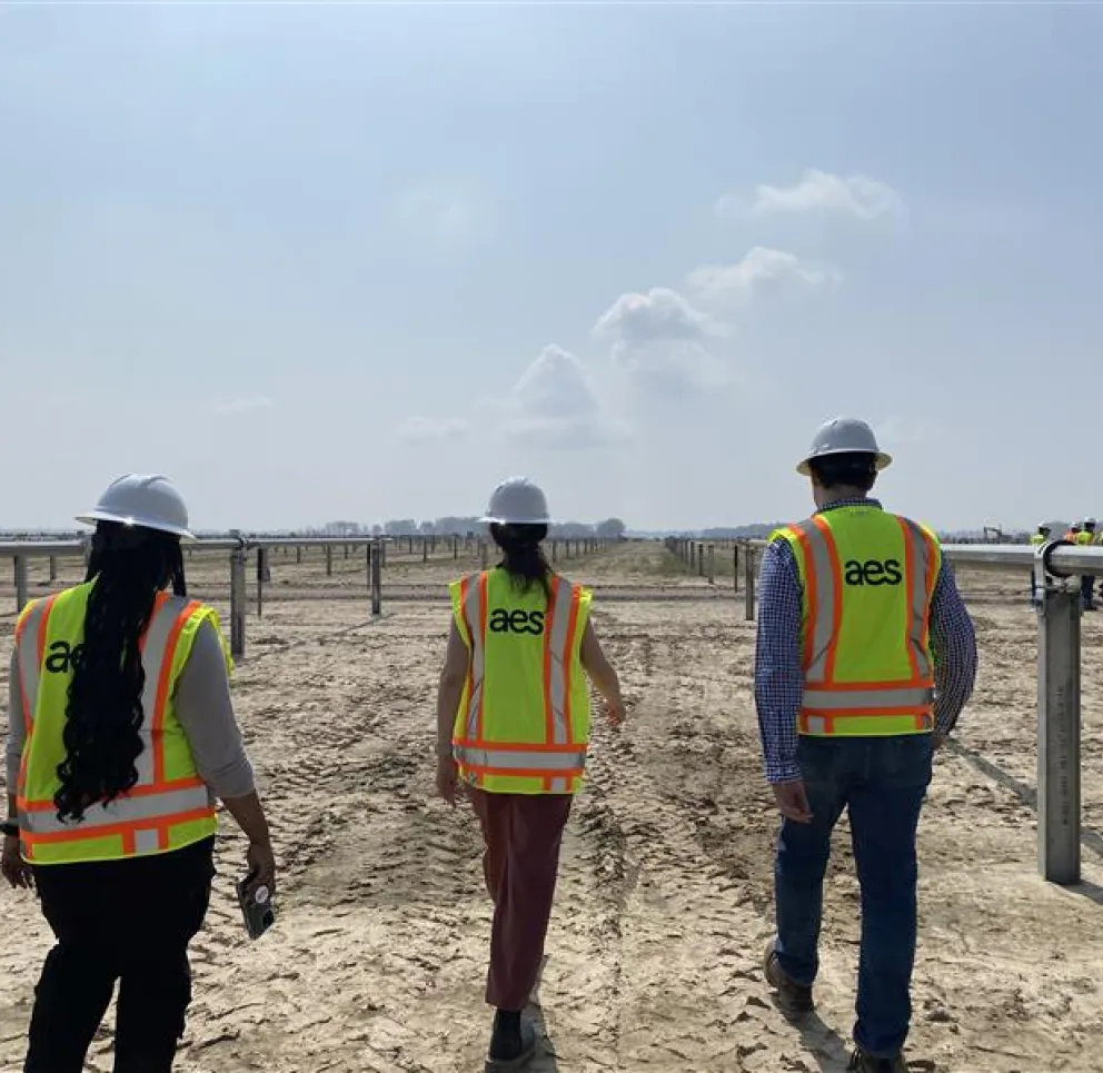 Three people wearing hard hats and high-visibility vests walk through a solar farm under a clear sky.
