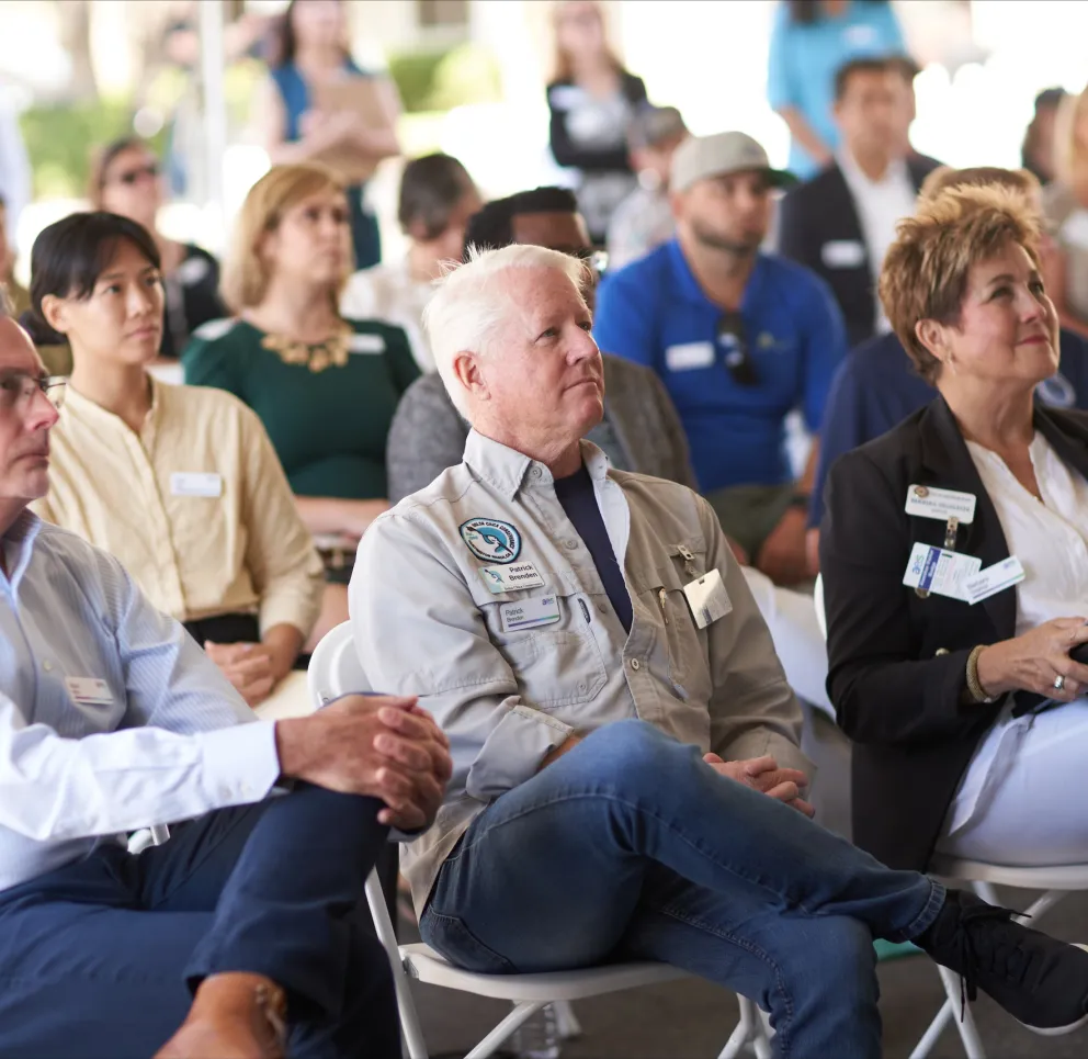 A group of people seated attentively in a conference setting, with name tags visible on their clothing. They are focused on a presentation or speaker out of frame.