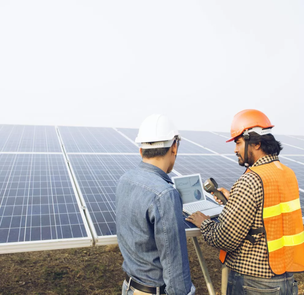 Two workers wearing hard hats and safety vests inspect solar panels. One holds a laptop showing graphs, while the other holds a device. They stand in a solar farm with rows of panels visible.