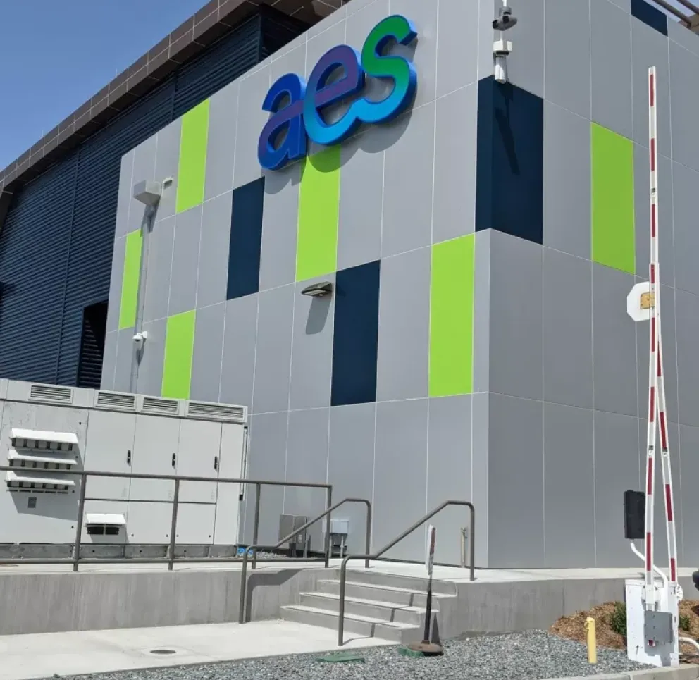 Modern building exterior with a geometric design featuring blue and green panels. A logo is displayed prominently on the wall. The area includes a security camera, railings, and a barrier gate.