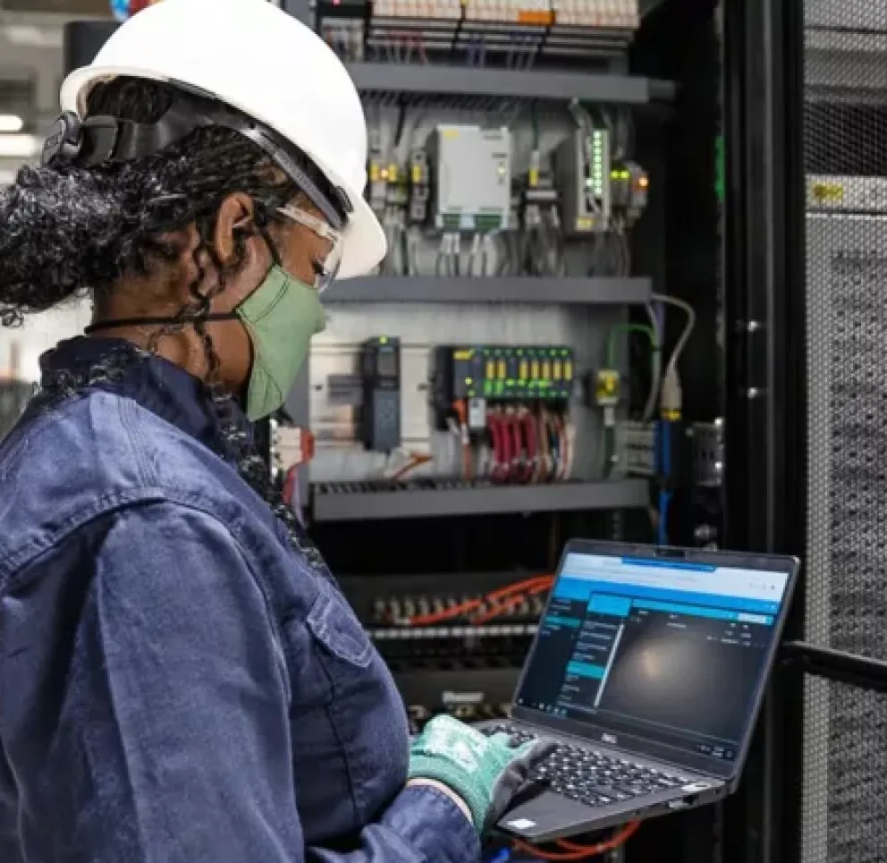 A person wearing a hard hat and mask works on a laptop in front of industrial equipment in a server room.