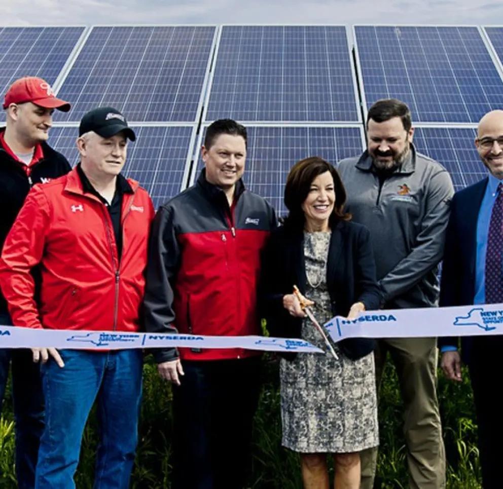 A group of people standing in front of solar panels, participating in a ribbon-cutting ceremony, with a woman holding scissors.