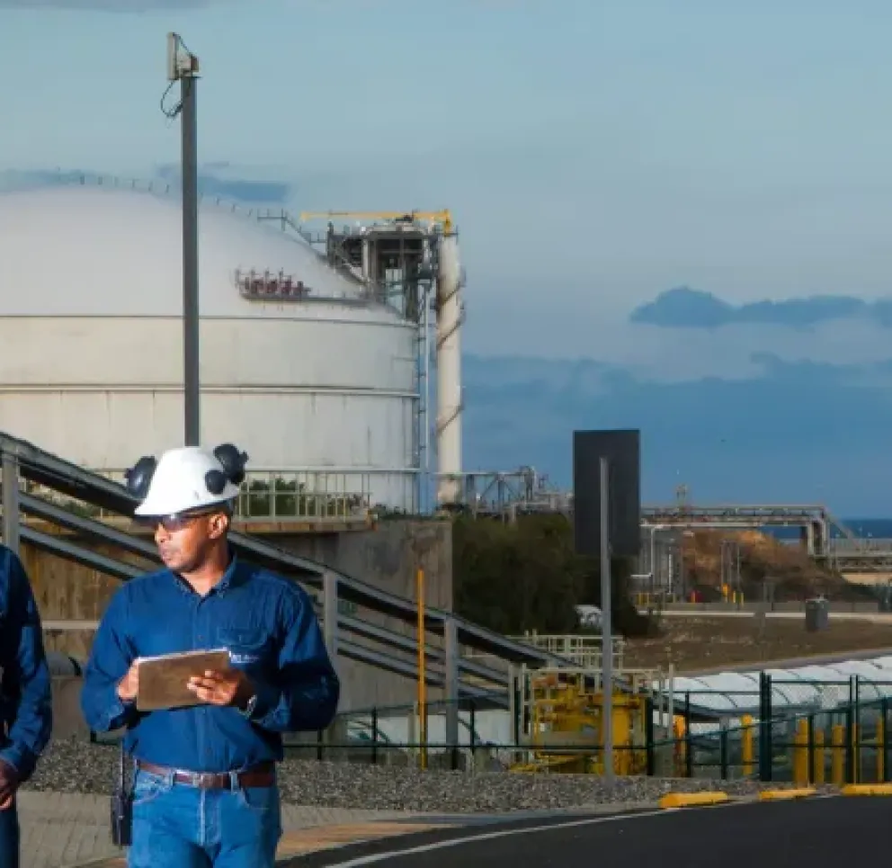 Two workers in blue uniforms and white hard hats walking and discussing near an industrial facility with large storage tanks.