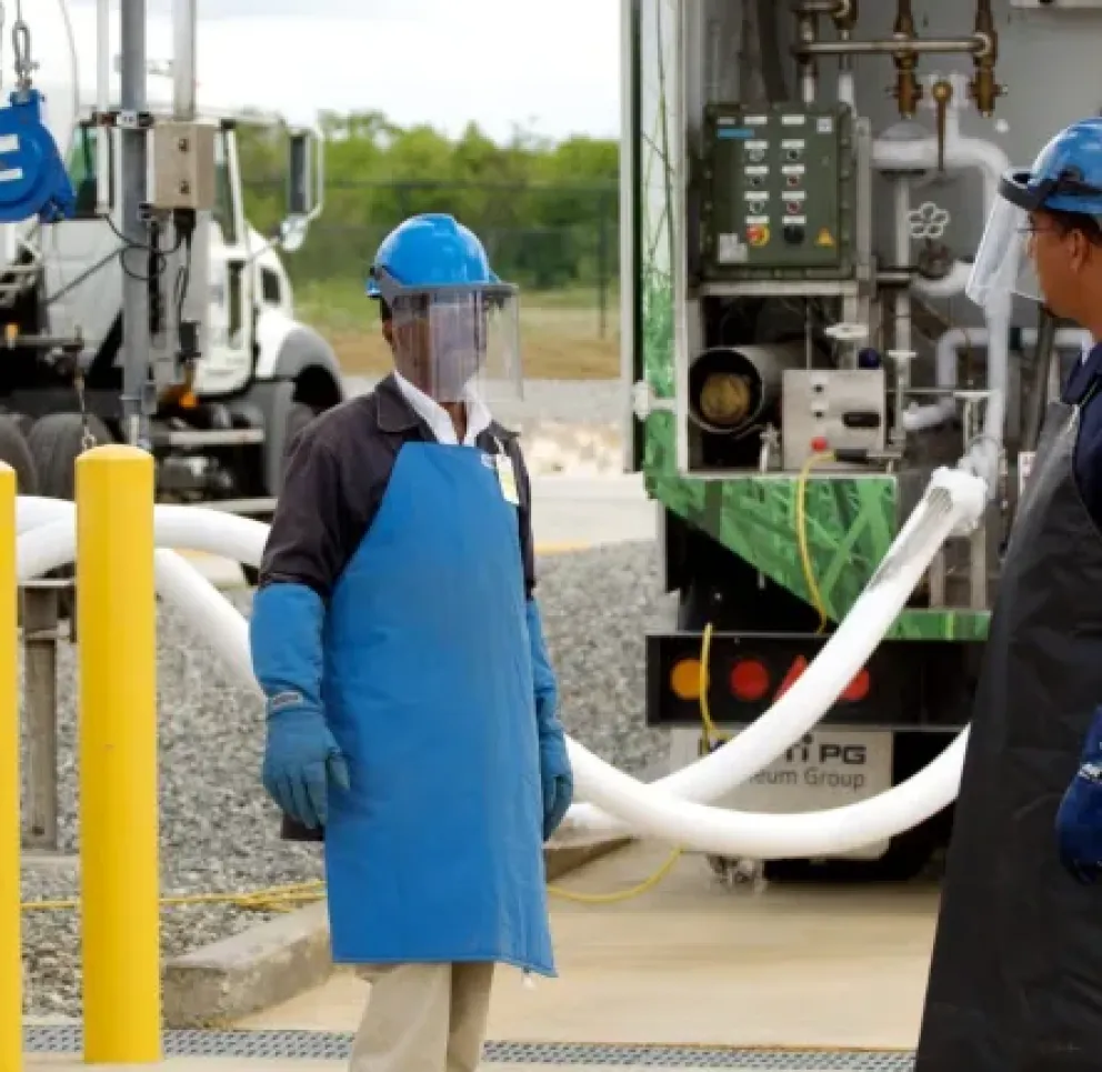Two workers wearing protective gear stand near a fuel truck, discussing. They are equipped with blue aprons, helmets, and face shields. The truck has hoses connected, and safety barriers are visible.