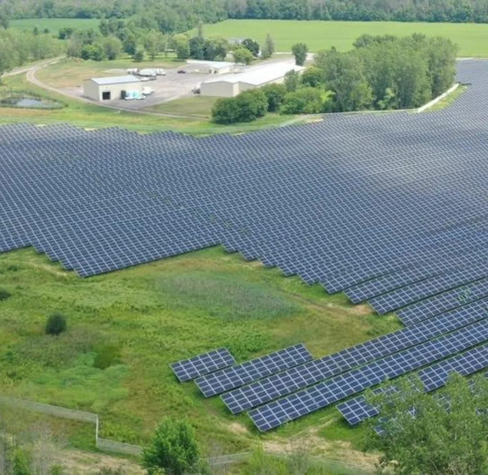Aerial view of a large solar farm with rows of solar panels covering a vast area surrounded by greenery and trees. A building complex is visible in the background.