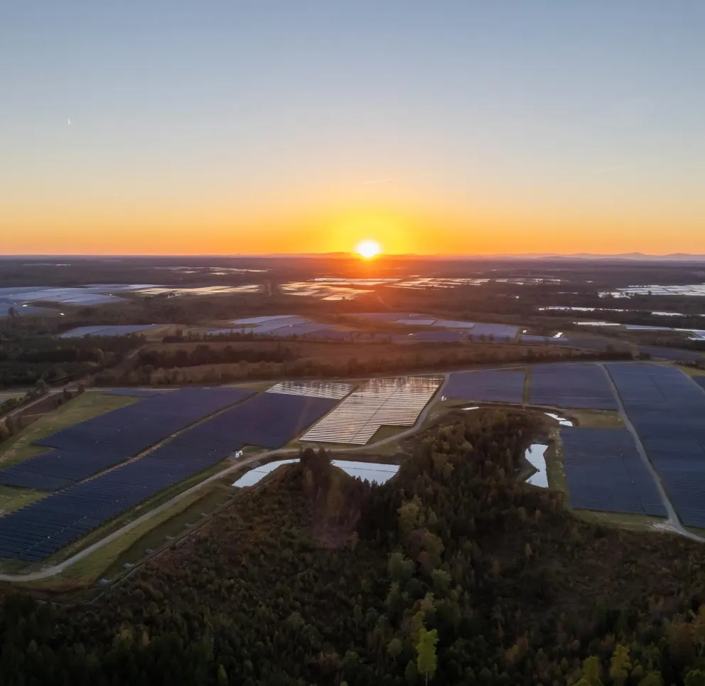 Aerial view of a large solar farm at sunset, with rows of solar panels reflecting the orange and purple hues of the sky. The landscape is surrounded by trees and distant hills under the colorful sky.