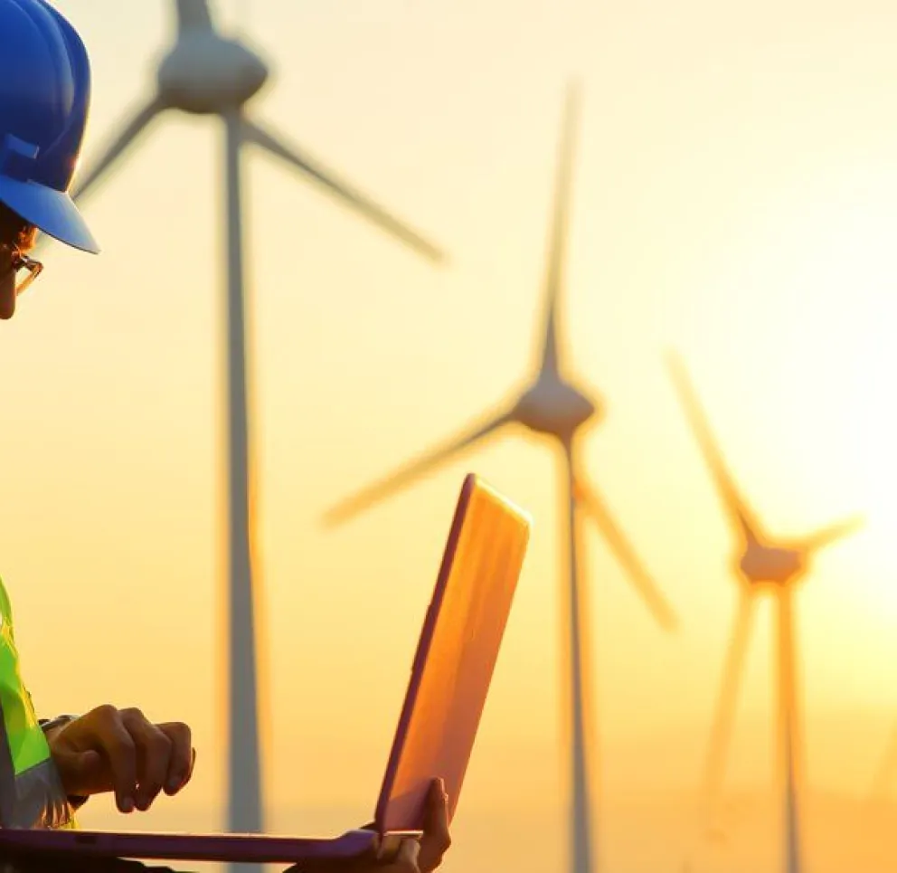 Two engineers wearing hard hats and reflective vests work on a laptop in front of wind turbines at sunset.