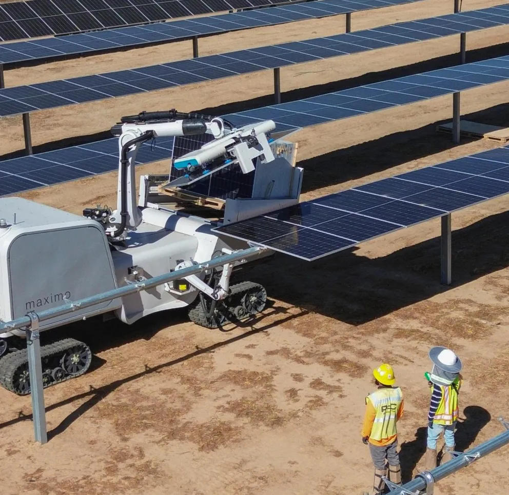 A robotic machine cleaning solar panels in a solar farm with two workers observing. The machine has a mechanical arm and is mounted on tracks, while the workers wear safety vests and helmets.