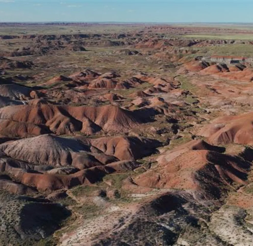 Aerial view of a vast desert landscape with rolling hills and colorful rock formations. The terrain is predominantly red and brown with patches of green vegetation scattered throughout. The sky is clear with a hint of blue on the horizon.