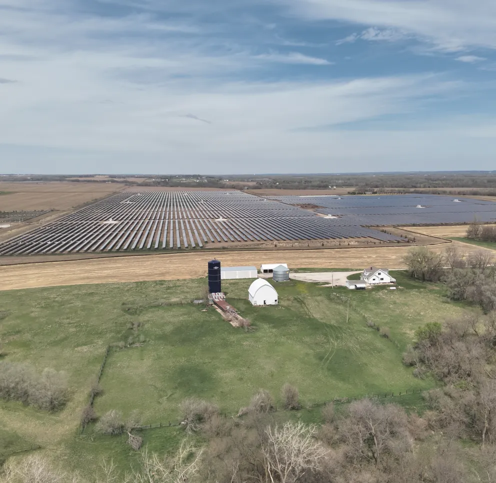 Aerial view of a large solar farm with rows of solar panels on a vast field. In the foreground, there is a small farm with a barn and silo surrounded by green fields and trees. The sky is partly cloudy.