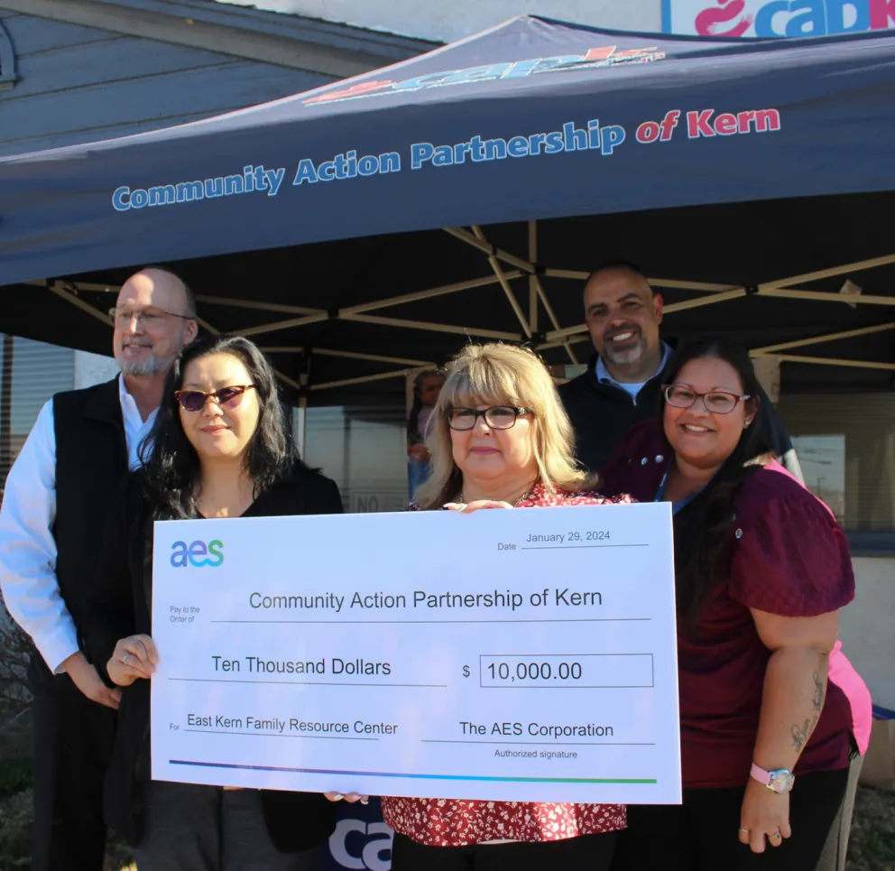 A group of five people stands under a canopy labeled 'Community Action Partnership of Kern.' They are holding a large check for ten thousand dollars from AES Corporation to the East Kern Family Resource Center. Two banners on either side display information about combating food insecurity and empowering youth and families.
