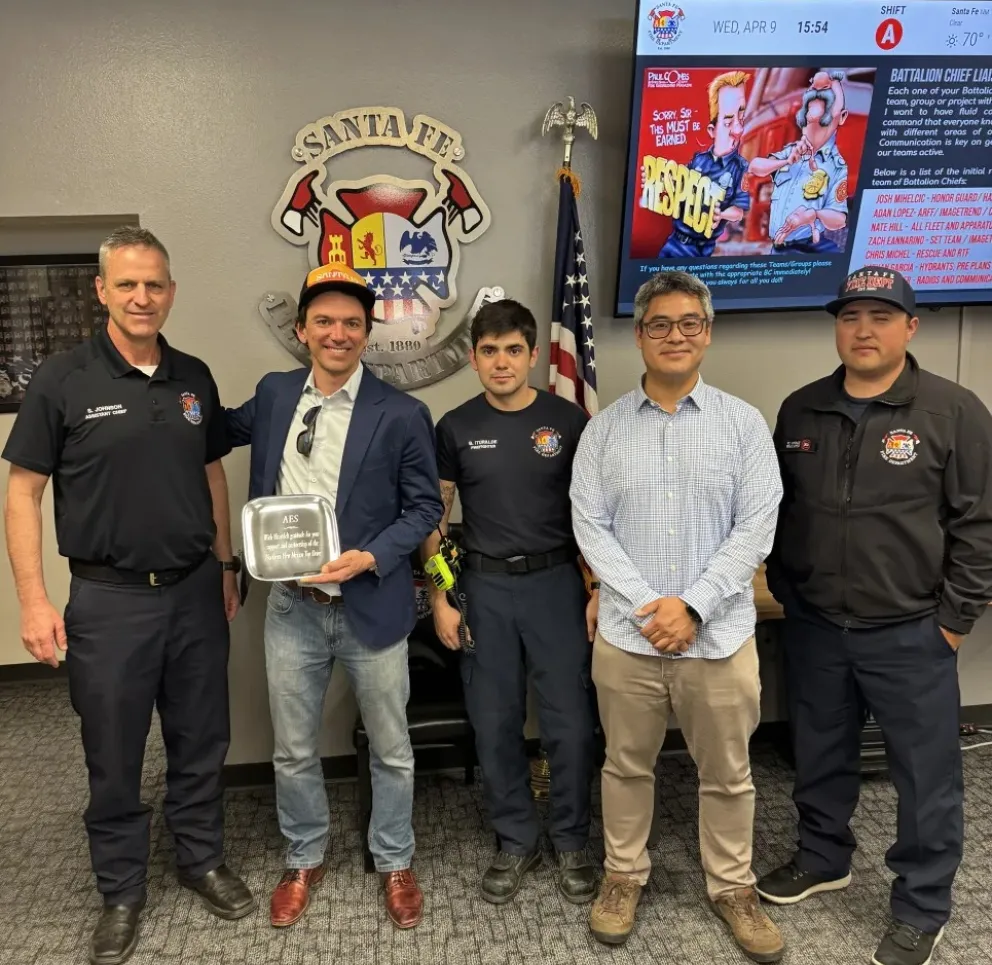 Five men standing in a room, with one holding a plaque. They are in front of a Santa Fe Fire Department emblem and a TV screen displaying information.