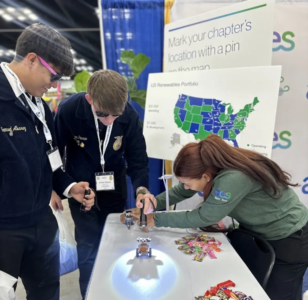Three people interact with small robots on a table at an event booth. Two young men in matching jackets observe as a woman in a green shirt demonstrates the robots. A map of the US renewables portfolio and a bowl of candy are visible on the table.