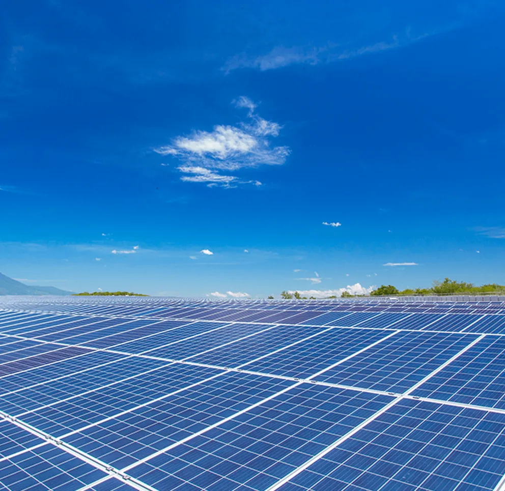 A large field of solar panels under a clear blue sky with a few clouds. The panels are aligned in rows, reflecting sunlight.