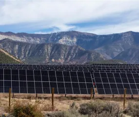 A field of solar panels is set against a backdrop of rugged mountains under a partly cloudy sky. The landscape has sparse vegetation and a fence surrounding the solar array.