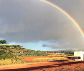 A vibrant rainbow arches across a cloudy sky above a rural landscape on Lawai with red dirt, green trees, grassy fields, and a white industrial battery storage container in the foreground.