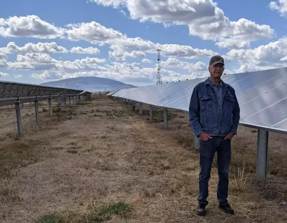 A man in a denim jacket and cap stands beside rows of solar panels in a field under a sky filled with fluffy clouds.