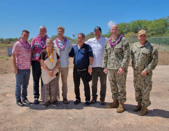 A group of eight people, including two in military uniforms, stand outdoors on a sunny day. Some wear leis and casual clothing. They are smiling, with trees and a clear blue sky in the background.