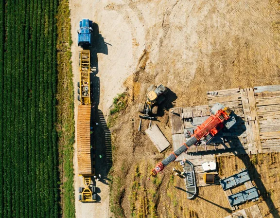 Aerial view of a construction site with a blue truck and a yellow trailer on a dirt path next to a green field. A red crane and construction materials are on the right side.