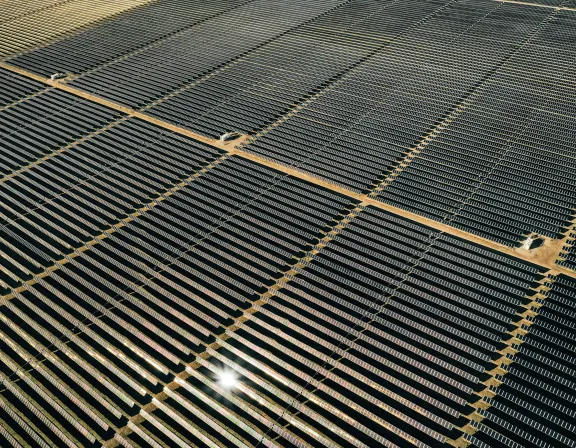 Aerial view of a large solar farm with rows of solar panels arranged in a grid pattern, reflecting sunlight.