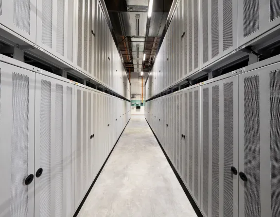 A narrow aisle in a data center with rows of tall, gray server racks on both sides. The floor is concrete, and the ceiling has exposed ducts and lighting fixtures.