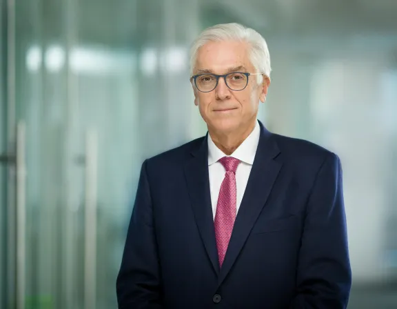 An older man with white hair and glasses wearing a navy suit and pink tie stands in a modern office with glass walls.