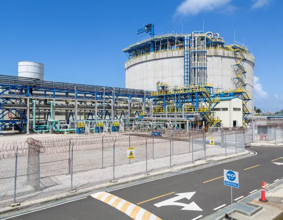 Industrial facility with large cylindrical structure, metal pipes, and safety signs, surrounded by a barbed wire fence under a clear blue sky.