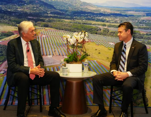 Two men in suits sit on chairs facing each other, with a table holding a potted orchid and water bottles between them. A large aerial photo of a solar farm is displayed in the background.