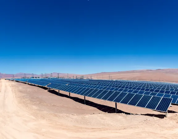 A vast solar farm in a desert landscape under a clear blue sky. Rows of solar panels are aligned on sandy terrain, with mountains visible in the distant background. A dirt road runs alongside the solar panels, bordered by a fence.