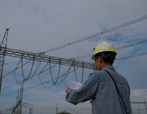 Engineer in a hard hat examining documents near a high-voltage power line, with a cloudy sky in the background.