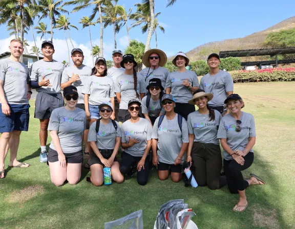 A group of people wearing matching gray t-shirts with 'AES Hawaii' printed on them, posing outdoors on a grassy area. Palm trees and a hill are visible in the background under a clear blue sky.