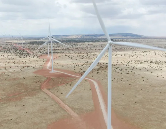 Aerial view of wind turbines on a vast, arid landscape with scattered vegetation and dirt roads under a cloudy sky.