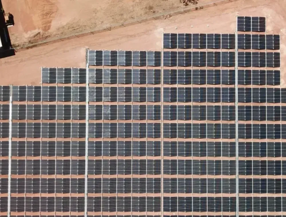 Aerial view of a large solar panel farm on a sandy terrain, with rows of dark solar panels arranged in a grid pattern.