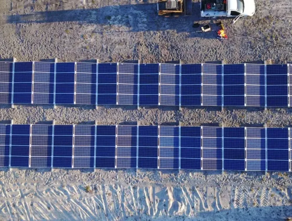 Aerial view of two rows of solar panels installed on sandy ground with a white vehicle and equipment nearby.