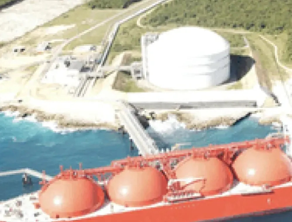Aerial view of a large red LNG tanker docked near a coastal storage facility with a white spherical tank and surrounding greenery.