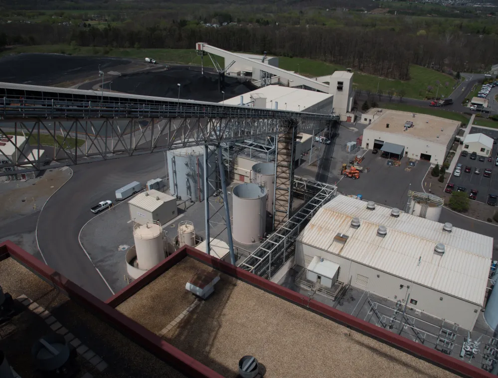 Aerial view of an industrial facility with large storage tanks, conveyor belts, and several buildings. Surrounding the facility are parking lots and green fields in the background.