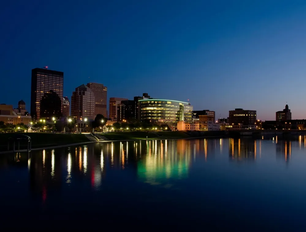 A city skyline at dusk with buildings illuminated by lights reflecting on a calm river. The sky is a deep blue transitioning to orange near the horizon.