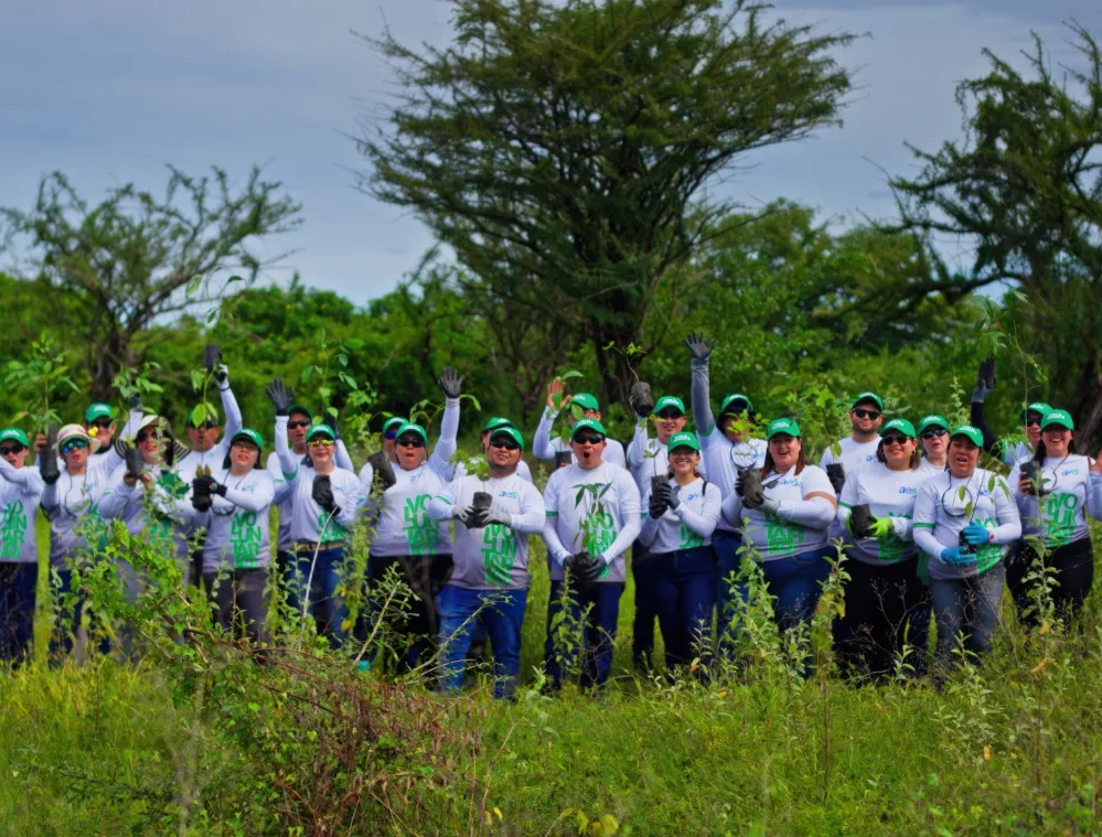 A group of people wearing matching white shirts and green caps stand in a field, holding small plants. They are smiling and appear to be participating in a tree planting activity. The background features lush green trees and a cloudy sky.