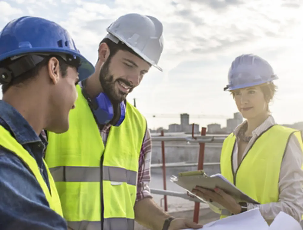 Three construction workers wearing hard hats and reflective vests review blueprints at a construction site. The sky is partly cloudy, and they appear to be discussing the plans.