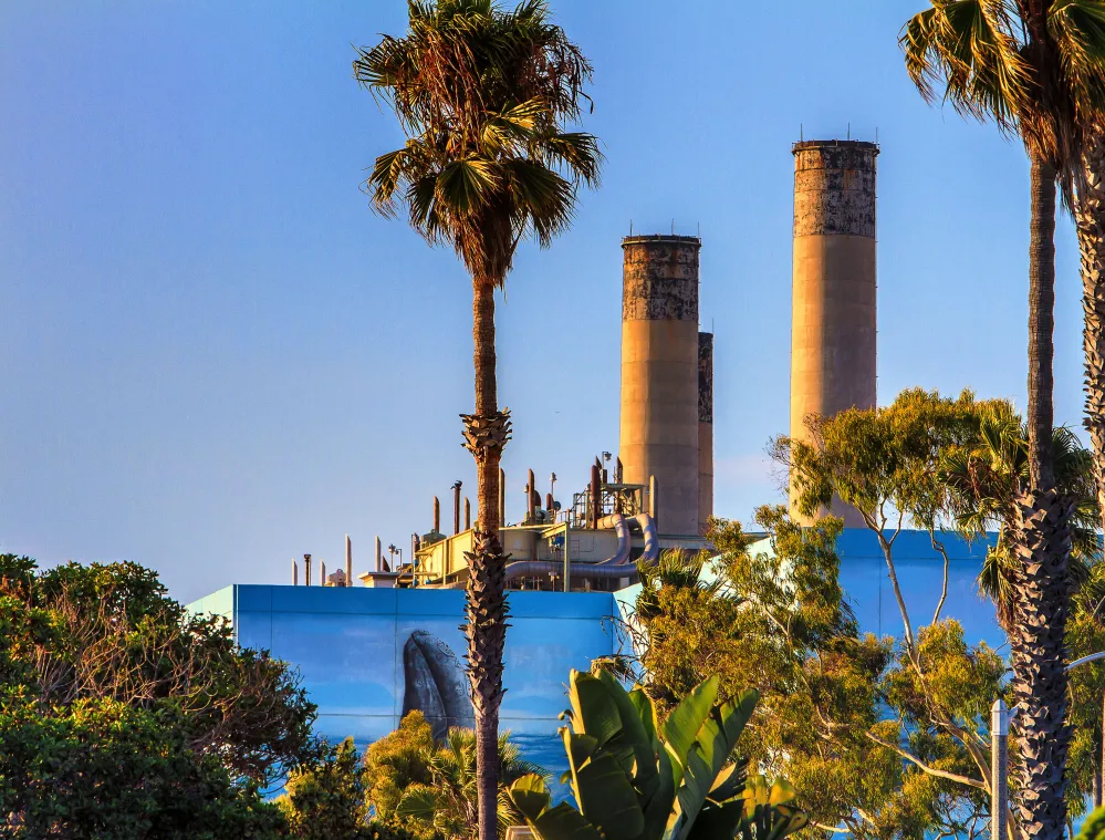 A power plant with two tall smokestacks surrounded by palm trees and greenery under a clear blue sky.