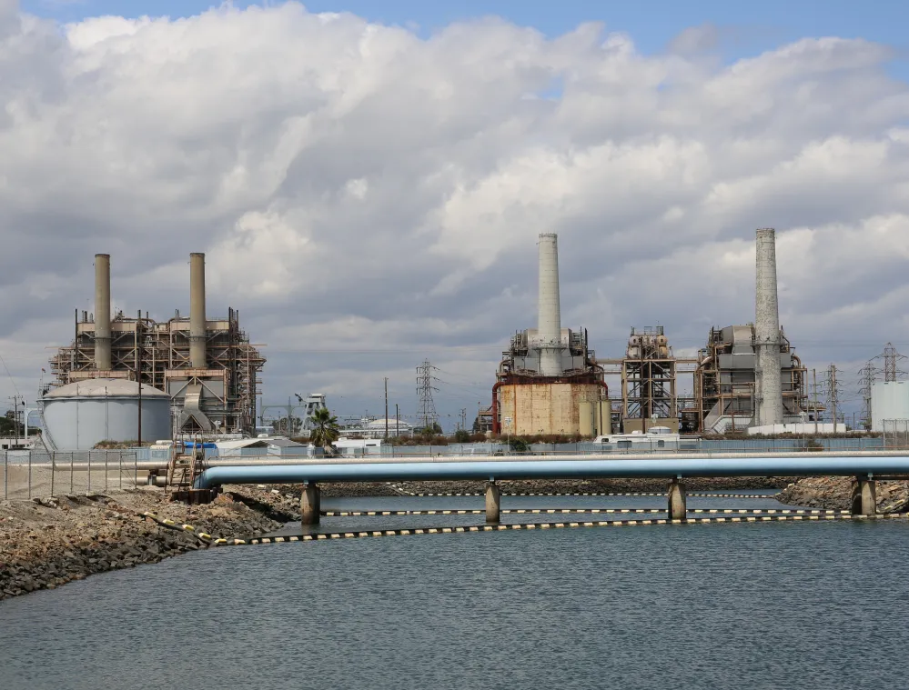 Industrial power plant with multiple tall chimneys and large cylindrical structures, set against a cloudy sky. A body of water is in the foreground with a blue pipeline crossing it.