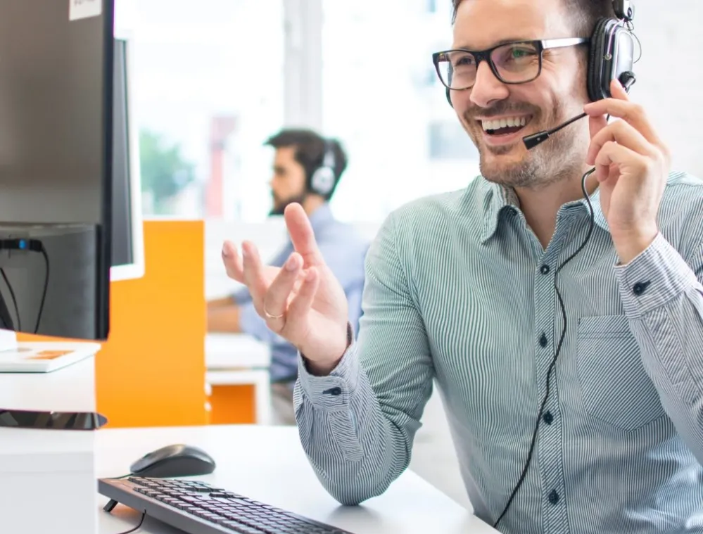 A man wearing glasses and a headset is sitting at a desk, smiling and gesturing with his hand. He is in front of a computer monitor and keyboard. In the background, another person wearing a headset is visible, slightly out of focus.
