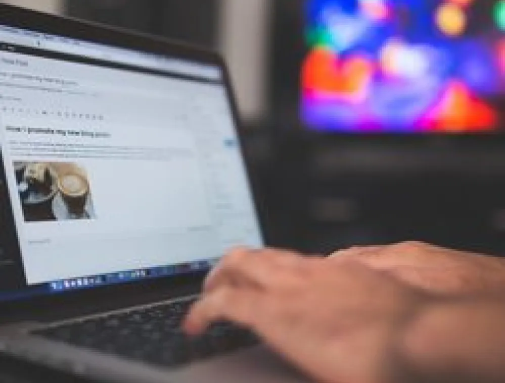 Person typing on a laptop with a blog post about coffee on the screen. A colorful, blurred background is visible.