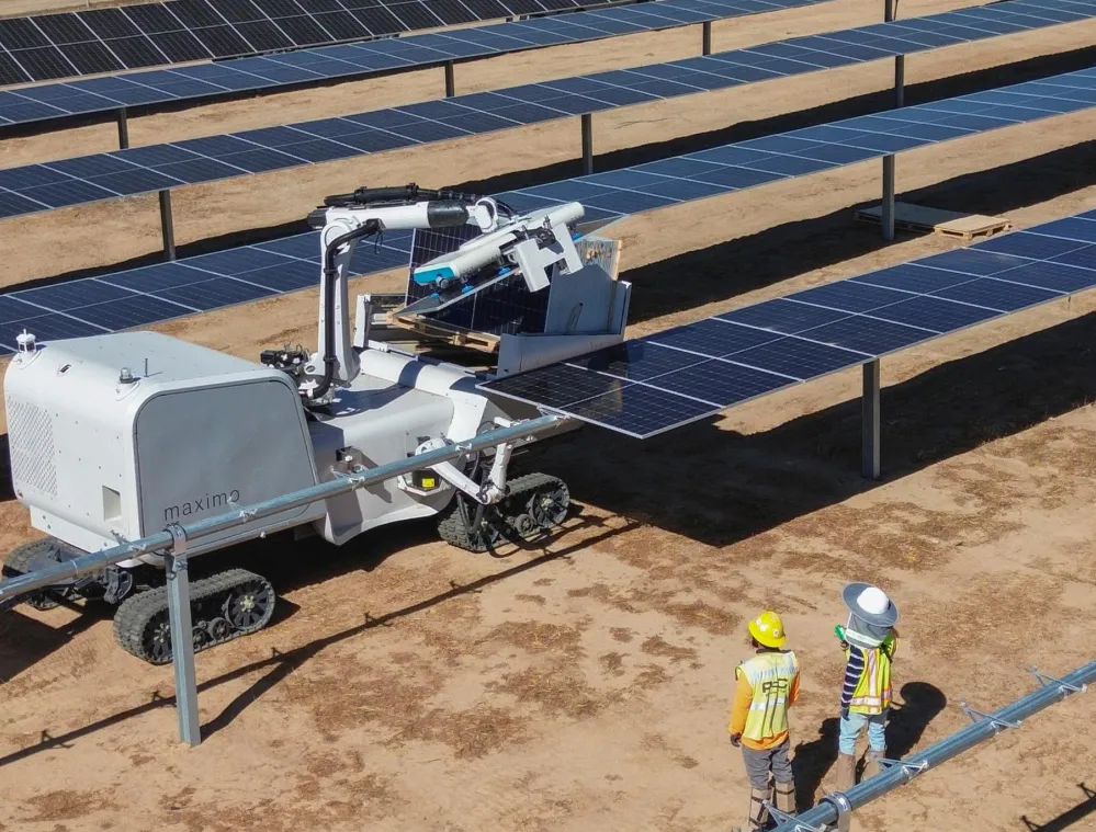 A robotic machine cleaning solar panels in a solar farm with two workers observing. The machine has a mechanical arm and is mounted on tracks, while the workers wear safety vests and helmets.