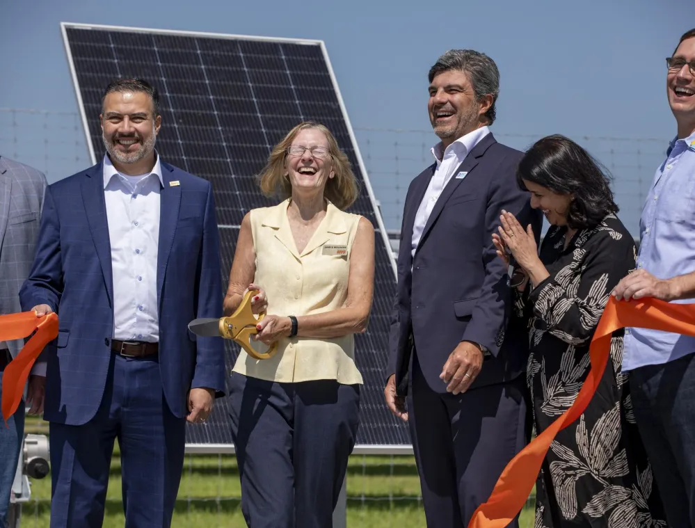 A group of people smiling and holding a large pair of scissors at a ribbon-cutting ceremony in front of a solar panel.