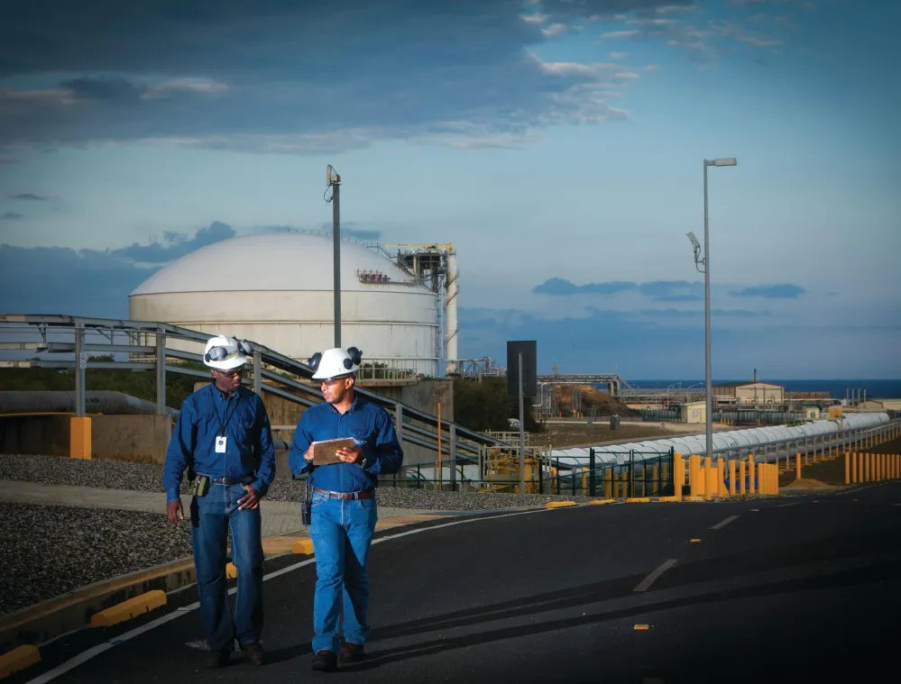 Two workers in blue uniforms and white hard hats walk along a road in an industrial area with large storage tanks and pipelines under a cloudy sky.