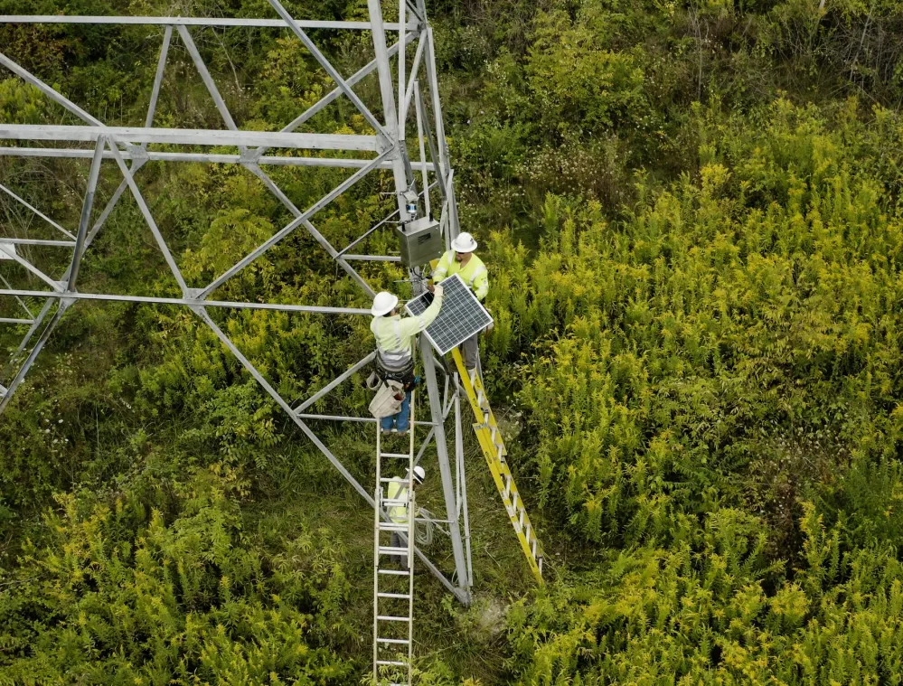 Two workers in safety gear installing a solar panel on a metal tower in a green, bushy area. One worker is on a ladder while the other is on the tower structure.